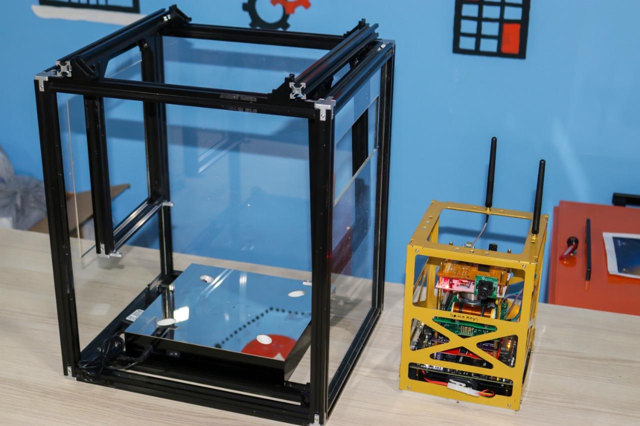 This image depicts technical items in a workshop-like environment at IVY STEM International School. On the left is an unassembled 3D printer framework, while on the right is a yellow-framed device with electronic components. The background features a blue wall with a decorative window element. Explore the hands-on learning opportunities at IVY STEM.